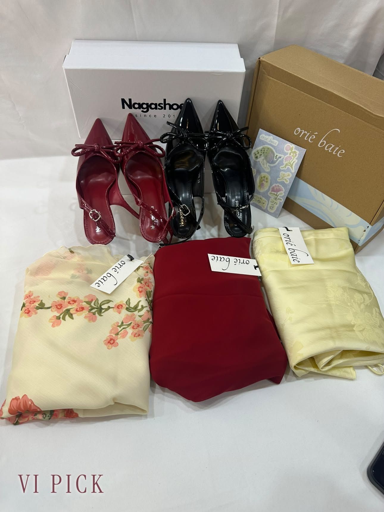 VI PICK QC flatlay for a US order: red and black NAGA Shoes bow slingback heels with the Nagashoes box, and three orié baie pieces in floral cream, red, and pale yellow with their brand tags